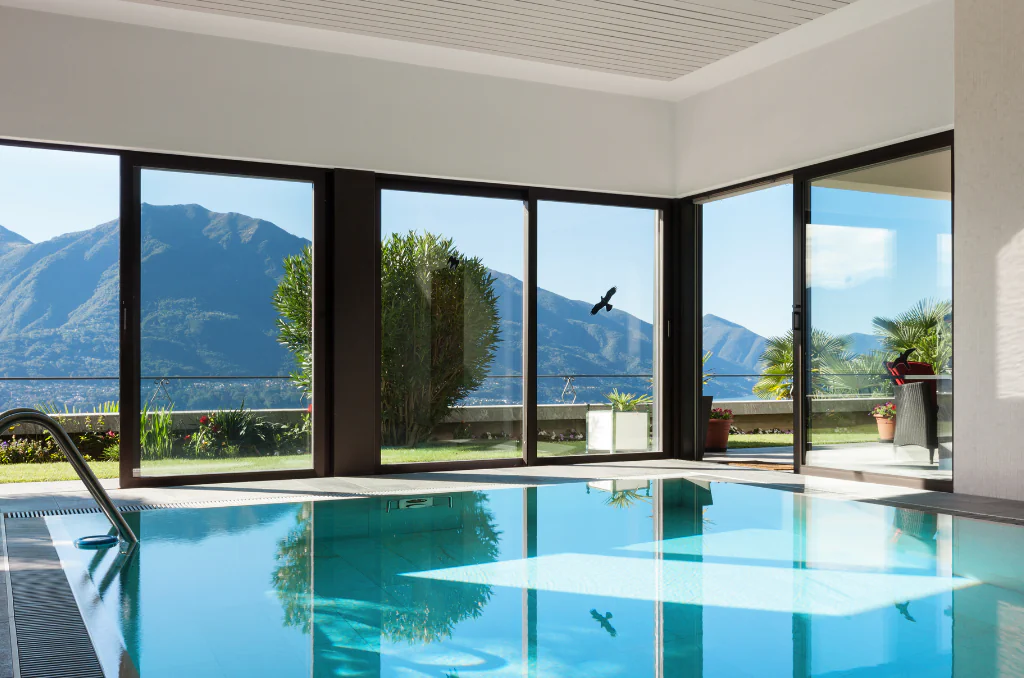 Ein Hallenbad mit klarem, blauem Wasser im Vordergrund, das die Fenster spiegelt. Große Glasschiebetüren bieten einen weiten Ausblick auf einen grünen Garten, Palmen und eine bergige Landschaft unter blauem Himmel.Ein Hallenbad mit klarem, blauem Wasser im Vordergrund, das die Fenster spiegelt. Große Glasschiebetüren bieten einen weiten Ausblick auf einen grünen Garten, Palmen und eine bergige Landschaft unter blauem Himmel. Ein Hallenbad mit klarem, blauem Wasser im Vordergrund, das die Fenster spiegelt. Große Glasschiebetüren bieten einen weiten Ausblick auf einen grünen Garten, Palmen und eine bergige Landschaft unter blauem Himmel.